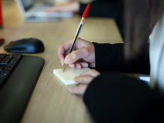 Primer plano de unas manos escribiendo algo en un post-it a boligrafo rojo.