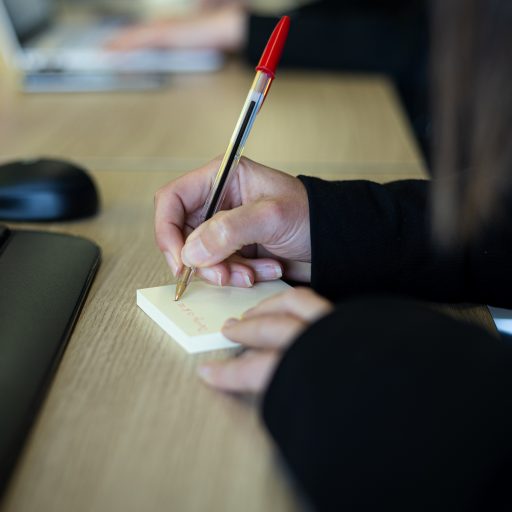 Primer plano de unas manos escribiendo algo en un post-it a boligrafo rojo.